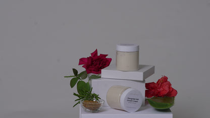 Two white cosmetic jars with cream-colored product displayed on white pedestals, styled with red flowers and small green plants against a plain gray background.