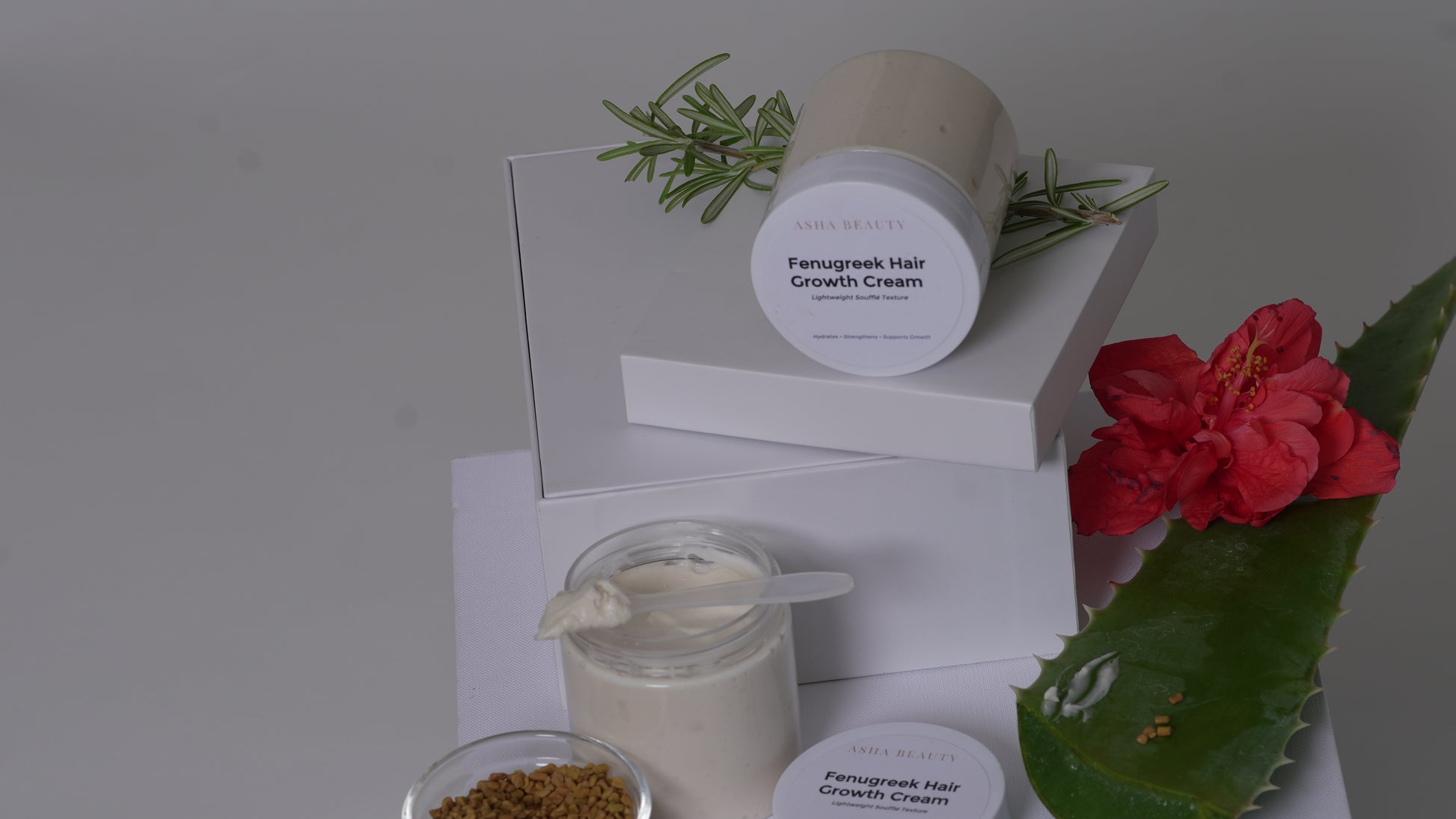 A styled product shot of a hair growth cream jar and its packaging boxes, decorated with rosemary sprigs, a red flower, and a green leaf on a neutral gray background.