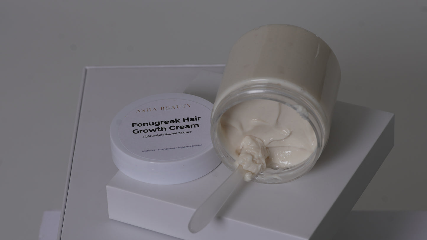A jar of Asha Belle Beauty Fenugreek Hair Growth Cream, with the cream visible in a clear glass container and its white lid resting beside it on a white display block.