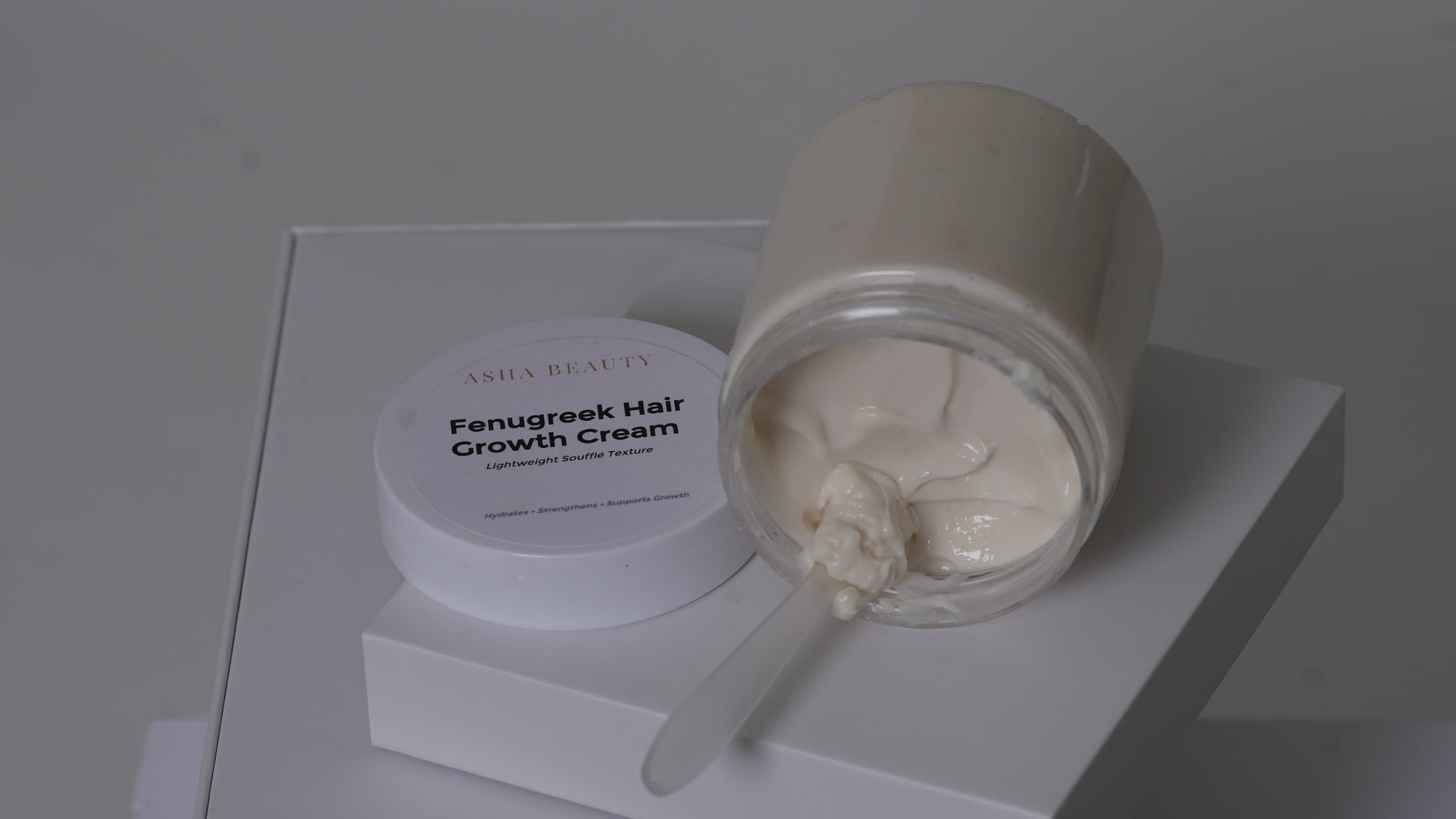 A jar of Asha Belle Beauty Fenugreek Hair Growth Cream, with the cream visible in a clear glass container and its white lid resting beside it on a white display block.