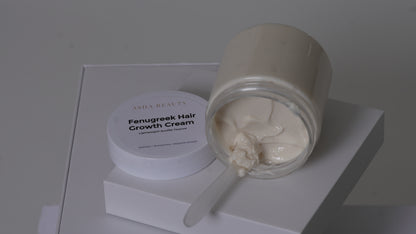 A jar of Asha Belle Beauty Fenugreek Hair Growth Cream, with the cream visible in a clear glass container and its white lid resting beside it on a white display block.