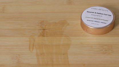 Asha Belle Beauty Nourish & Define Curl Gel jar on light wood surface next to wet glossy section showing smoothing and defining effect